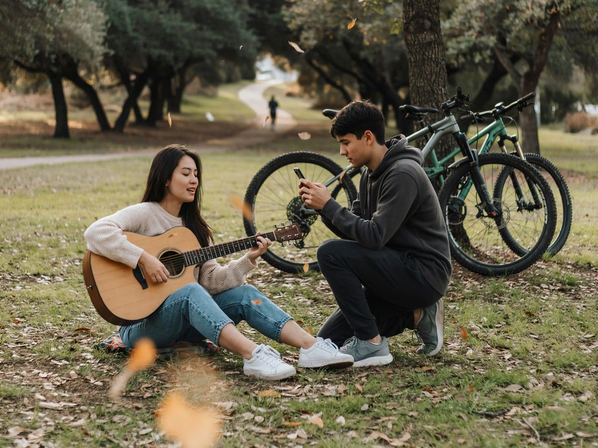 Una chica toca la guitarra y un chico mira su celular en un parque, junto a dos bicicletas
